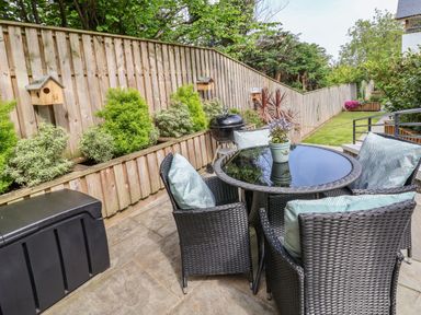 A patio with a round glass table surrounded by four wicker chairs with cushions next to a wooden fence with birdhouses and shrubs at The Nest in Prestatyn