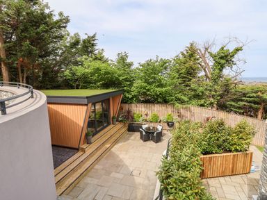 An outdoor patio area with a small wooden building with a grass roof surrounded by trees and plants at The Nest in Prestatyn