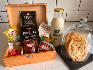 A wooden box with various Welsh tea and coffee packets a bottle of milk and cookies under a glass dome on a counter at The Nest in Prestatyn