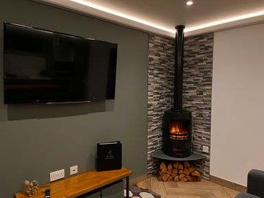 A room with a wall-mounted television a wood stove with stacked firewood and a wooden table with decorative items and a guest information folder at The Nest in Prestatyn