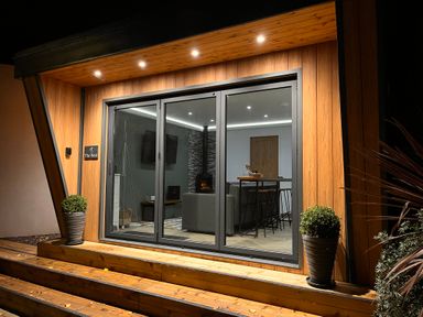 A wooden porch with potted plants and large glass sliding doors showing a living area with a fireplace at The Nest in Prestatyn