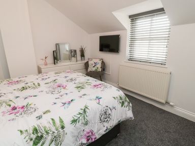 A bedroom with a bed covered with floral bedding a dresser with a mirror a chair with a decorative pillow a wall-mounted TV a window with blinds and a radiator at Lavenna in Alnwick
