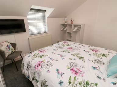 A bedroom with a bed covered in floral bedding a chair with a cushion a wall-mounted TV and an open wardrobe at Lavenna in Alnwick