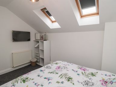A bedroom with a bed with floral bedding a white shelving unit a wall mounted television and two skylight windows