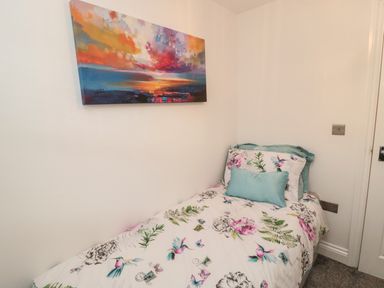 A single bed with floral and bird patterned bedding and a teal pillow in a small room with white walls and a colorful abstract painting on the wall at Lavenna in Alnwick