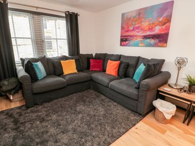 A living room with a dark gray sectional sofa with colorful pillows a side table with a lamp and plant and a painting on the wall at Lavenna in Alnwick