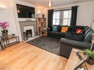 A living room with a dark sofa, fireplace, TV mounted on the wall, window with blinds and black curtains at Lavenna in Alnwick