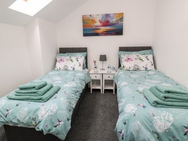 A bedroom with two single beds with blue floral bedding and towels on the beds a nightstand between the beds with a lamp and water bottles and a colorful painting on the wall at Lavenna in Alnwick