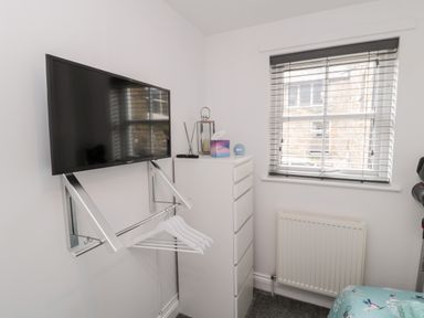 A small room with a wall-mounted TV above a hanging rail with hangers a white tall dresser a window with blinds and a radiator below at Lavenna in Alnwick