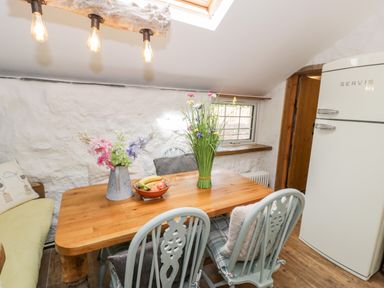 A kitchen dining area with a wooden table flowers in vases a bowl of fruit pale blue chairs a cream fridge and a cushioned bench at Gull Cottage Hermon near Newborough