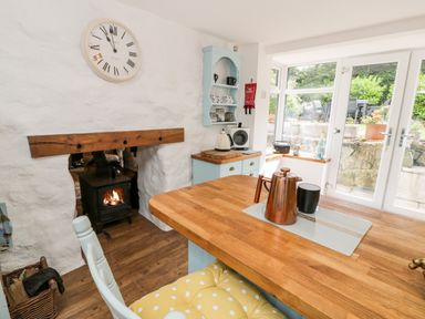 A kitchen with a wood stove, a wooden table with a copper teapot and black cup and doors opening to a garden at Gull Cottage in Hermon near Newborough