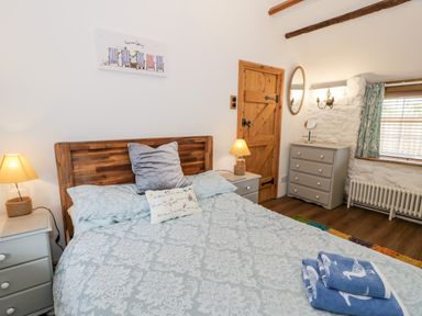 A bedroom with a bed with patterned bedding two bedside tables with lamps a wooden door a chest of drawers a mirror and a window with curtains at Gull Cottage Hermon near Newborough