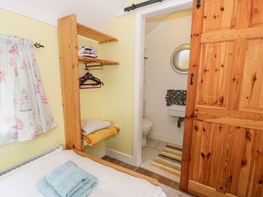 A bedroom with a bed and folded towels on it a wooden shelf with towels hangers and an iron next to a bathroom with a toilet and sink behind a sliding wooden door at Gull Cottage in Hermon near Newborough