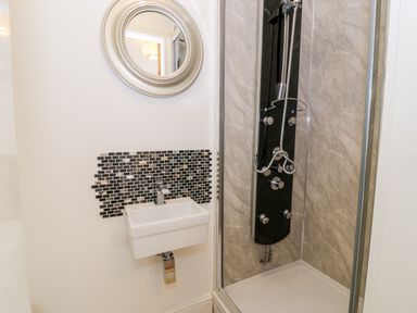 A bathroom with a wall-mounted sink below a round mirror and a shower enclosure with a black shower panel at Gull Cottage in Hermon near Newborough
