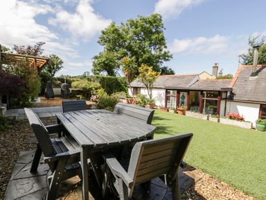 An outdoor patio area with a wooden table and chairs next to a lawn and small buildings at Gull Cottage in Hermon near Newborough