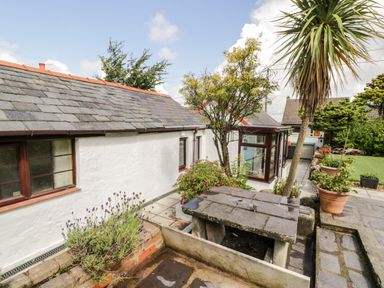 An outdoor patio area with stone table and benches surrounded by plants next to a white house with tiled roof at Gull Cottage in Hermon near Newborough