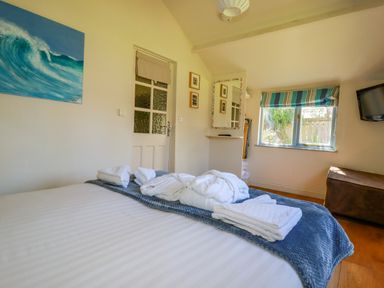 A bedroom with a bed covered with white linens and towels a door a window with striped blinds a wall-mounted tv and a painting of a wave at Abalone in Bude