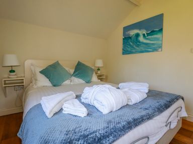 A bedroom with a bed made with white and blue bedding with white towels and robes on the bed and a painting of a wave on the wall at Abalone in Bude