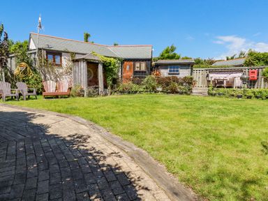 A backyard with a grassy lawn a paved pathway wooden garden chairs small sheds and a patio area at Seashells Atlantic Highway near Bude