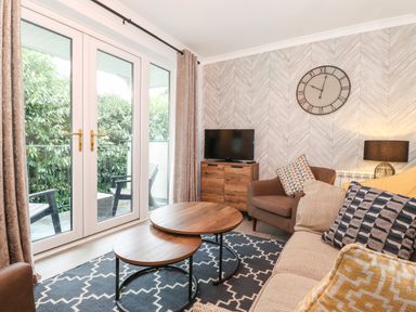A living room with a sofa, armchair, nested coffee tables, a TV on a wooden cabinet, wall clock, lamp, and patio doors opening to outside at Bluebell Hill in Bowness-On-Windermere