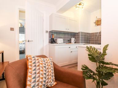 A small kitchen with white cabinets and gray tiled backsplash next to a brown armchair with a patterned cushion and a green plant at Bluebell Hill in Bowness-On-Windermere