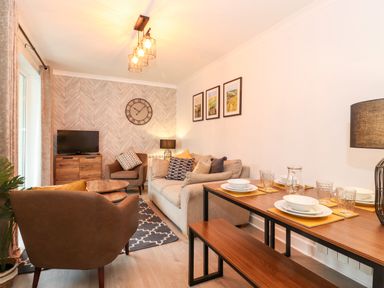 A living room with a beige sofa brown armchairs a wooden dining table with tableware and a television on a cabinet at Bluebell Hill in Bowness-On-Windermere