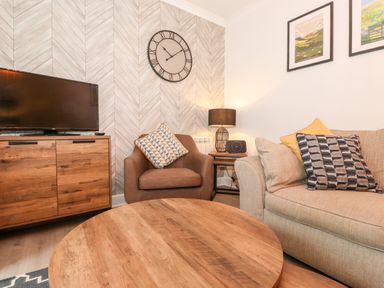 A living room with a wooden coffee table a beige sofa and a brown armchair with cushions a wooden media cabinet a flat screen tv a wall clock and framed pictures at Bluebell Hill in Bowness-On-Windermere