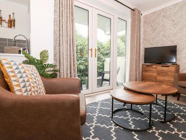 A living room with two brown armchairs a wooden cabinet with a TV two nested coffee tables a dark patterned rug and glass doors with curtains at Bluebell Hill in Bowness-On-Windermere