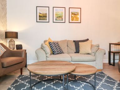A living room with a beige sofa and pillows a brown armchair wooden coffee tables and framed pictures on the wall at Bluebell Hill in Bowness-On-Windermere