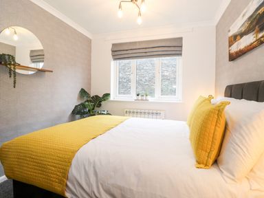 A bedroom with a bed with yellow pillows and blanket a plant a round mirror on the wall and a window with gray blinds at Bluebell Hill in Bowness-On-Windermere