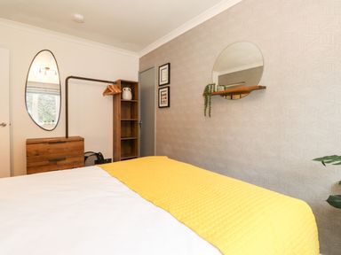 A bedroom with a bed covered in white and yellow bedding a wooden dresser a tall open shelf with hangers a round mirror and a wall shelf with a plant at Bluebell Hill in Bowness-On-Windermere