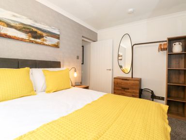 A bedroom with a bed with yellow bedding a wooden chest of drawers an oval mirror and an open wardrobe at Bluebell Hill in Bowness-On-Windermere