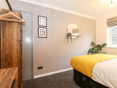 A bedroom with a wooden wardrobe, a bed with a yellow blanket, a round mirror with a shelf and plant, and two framed pictures on the wall at Bluebell Hill in Bowness-On-Windermere