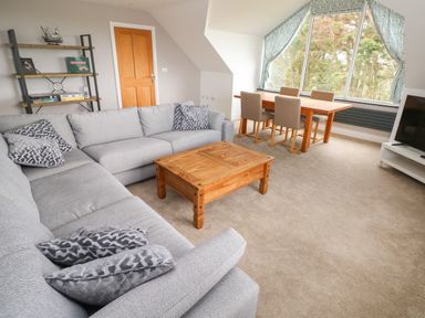 A living room with a gray sectional sofa and wooden coffee table near a dining table with four chairs by a large window at Landfall in St Twynnells near Pembroke