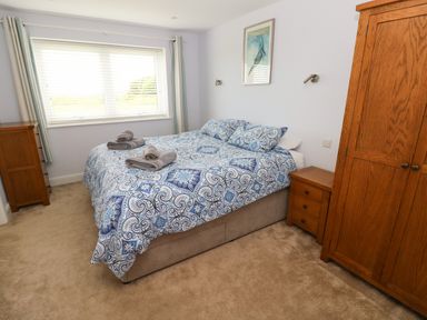 A bedroom with a bed covered in blue patterned bedding two folded towels on it wooden wardrobe wooden chest of drawers and window with blinds at Landfall in St Twynnells near Pembroke