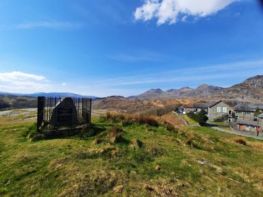 A grassy hill with a fenced monument and buildings in the background surrounded by mountains at Bridgeway House in Blaenau Ffestiniog