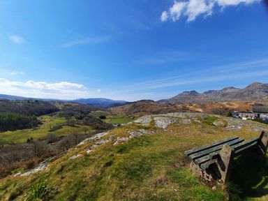 A bench on a grassy hill overlooking a valley with trees and mountains under a blue sky at Bridgeway House in Blaenau Ffestiniog