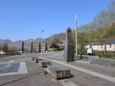 A paved outdoor area with benches stone monument flagpole and triangular towers with mountains in the background at Bridgeway House in Blaenau Ffestiniog