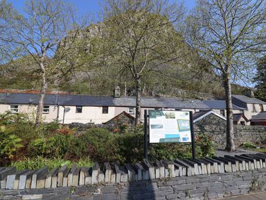 A row of stone houses with a slate fence and informational sign in front with trees and a mountain behind at Bridgeway House in Blaenau Ffestiniog
