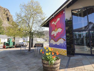 A colorful mural with birds on a building next to a tree and flower pot with tulips on a paved street with a green toy train in the background at Bridgeway House in Blaenau Ffestiniog