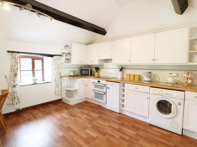 A kitchen with white cabinets appliances and a wooden floor with a window with floral curtains at The Corn Tallet in Westleigh near Bideford