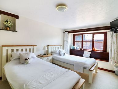A bedroom with two single beds a wooden nightstand with a lamp a window with curtains and a small television mounted at The Corn Tallet in Westleigh near Bideford