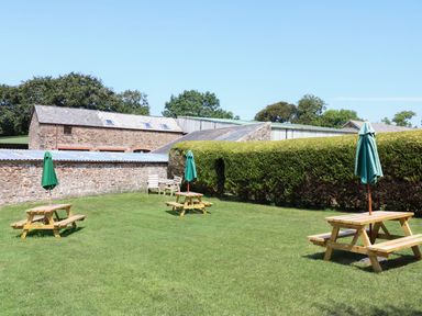 An outdoor grassy area with wooden picnic tables and umbrellas near stone and metal farm buildings at The Corn Tallet in Westleigh near Bideford