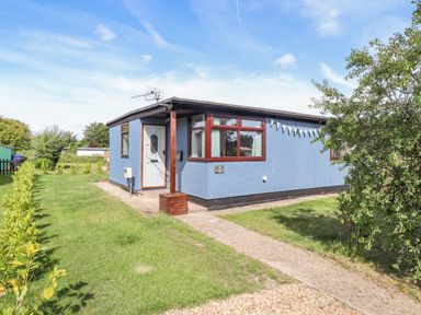 A blue single-story building with a white door and windows surrounded by grass and small bushes at Rollin Dunes in Humberston Lincolnshire