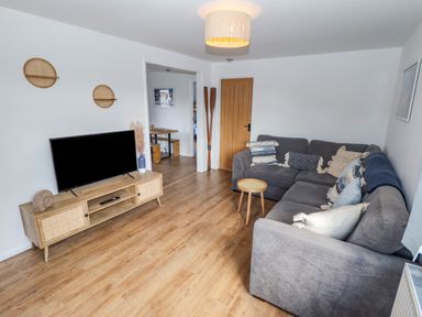A living room with a grey sofa, wooden TV stand with a TV, wall shelves, and a wooden door at Rollin Dunes in Humberston Lincolnshire