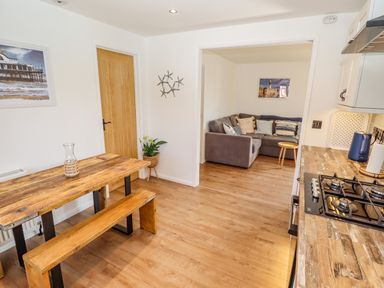 A kitchen with wooden table and bench along with a view into a living room with gray sofa and cushions at Rollin Dunes in Humberston Lincolnshire