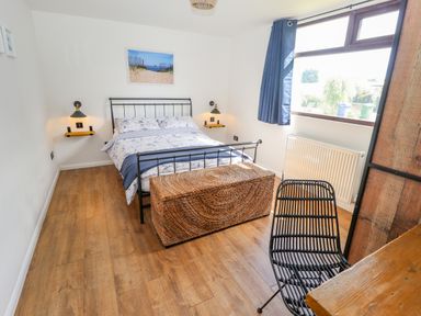 A bedroom with a double bed two wall lamps a wicker storage chest a black chair and a window with blue curtains at Rollin Dunes in Humberston Lincolnshire