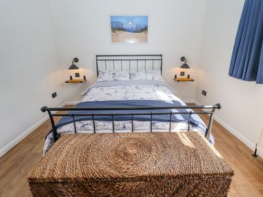 A bedroom with a metal bed, patterned bedding, wall lamps, a wicker chest, and a beach picture above the bed at Rollin Dunes in Humberston Lincolnshire