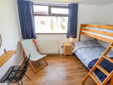 A bedroom with a wooden bunk bed a small bedside table with a lamp a blue checkered deck chair a black chair wooden flooring and a large window with blue curtains at Rollin Dunes in Humberston Lincolnshire