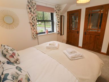 A bedroom with a bed with folded towels floral pillows a window and wooden double doors at Wood Brook Cottage in Crowan